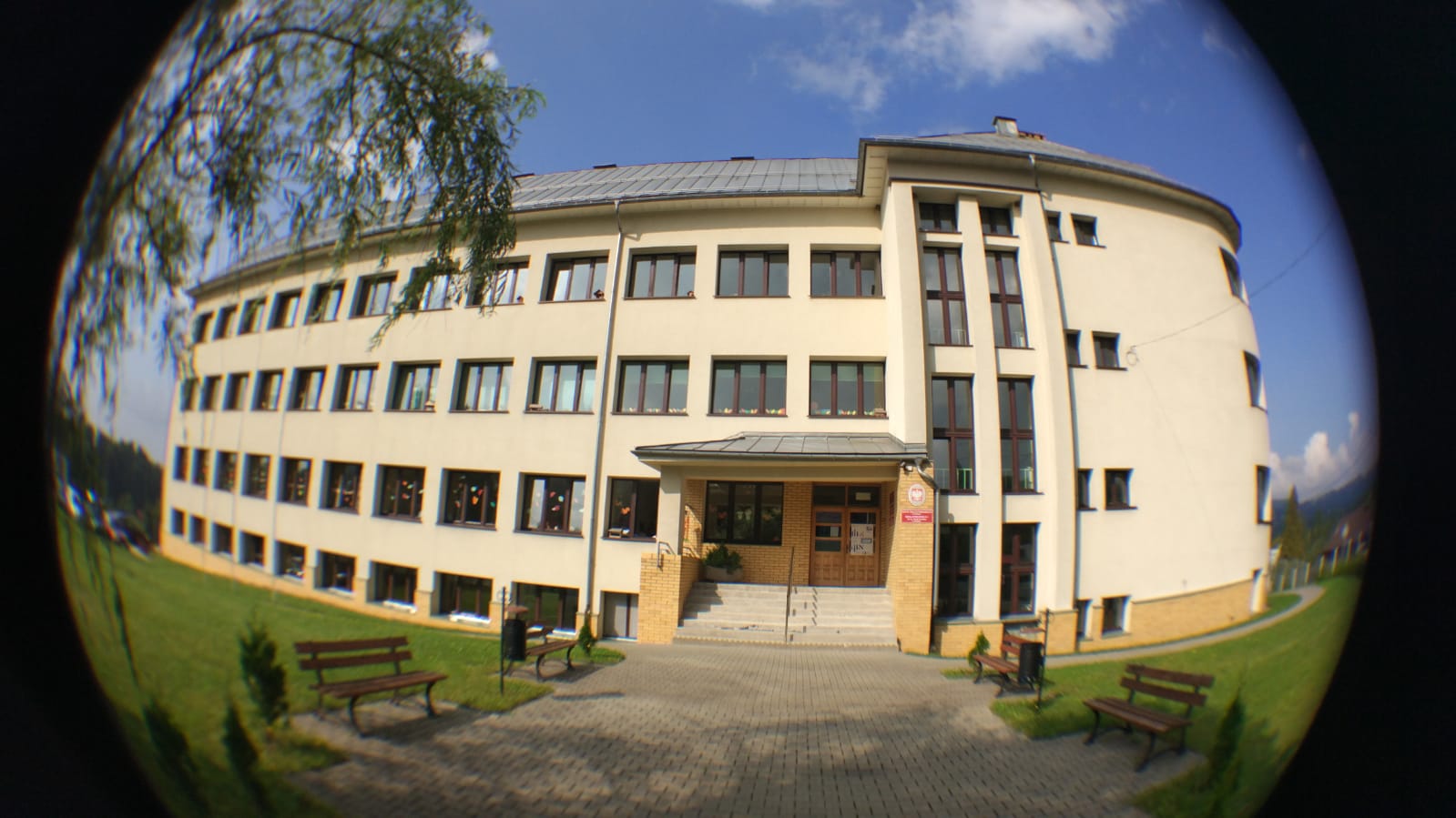 Czterokondygnacyjny budynek o jasnej elewacji z ciemnymi oknami. Wejście główne z zadaszeniem i schodami, otoczone ceglanym wykończeniem. Przed budynkiem chodnik z ławkami i trawnikiem.